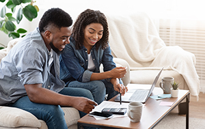 A man and a woman sitting on a couch with a laptop, bills and a calculator. 